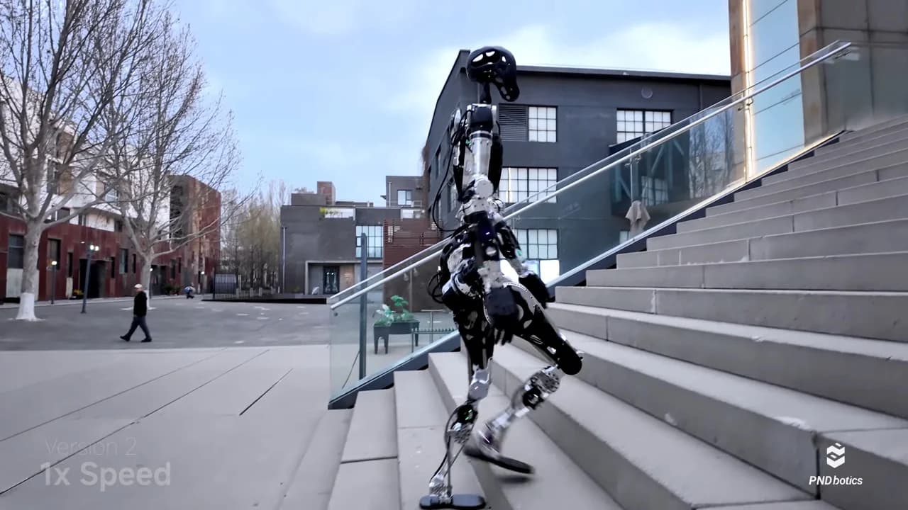 A silver and black PNDbotics Adam humanoid robot, characterized by its perforated torso and head, is shown from behind walking up a wide set of outdoor concrete stairs. The robot is captured mid-stride in an urban setting with modern gray buildings and a glass railing in the background. The bottom corners of the image display the PNDbotics logo and text identifying the footage as "Version 2" at "1x Speed."