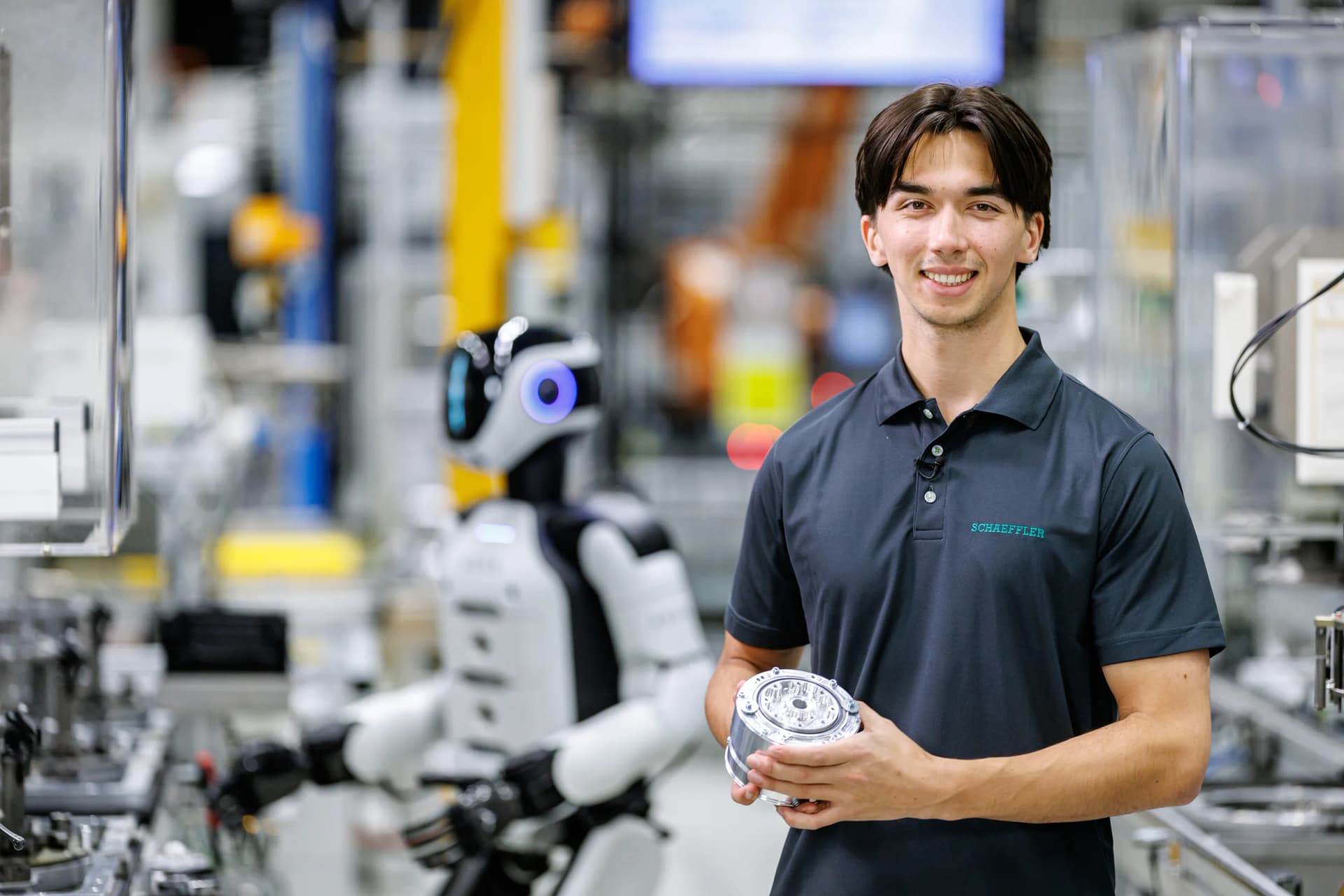 A Schaeffler employee stands in a factory setting holding a metallic planetary gear actuator in both hands. A white and black humanoid robot stands out of focus in the background.
