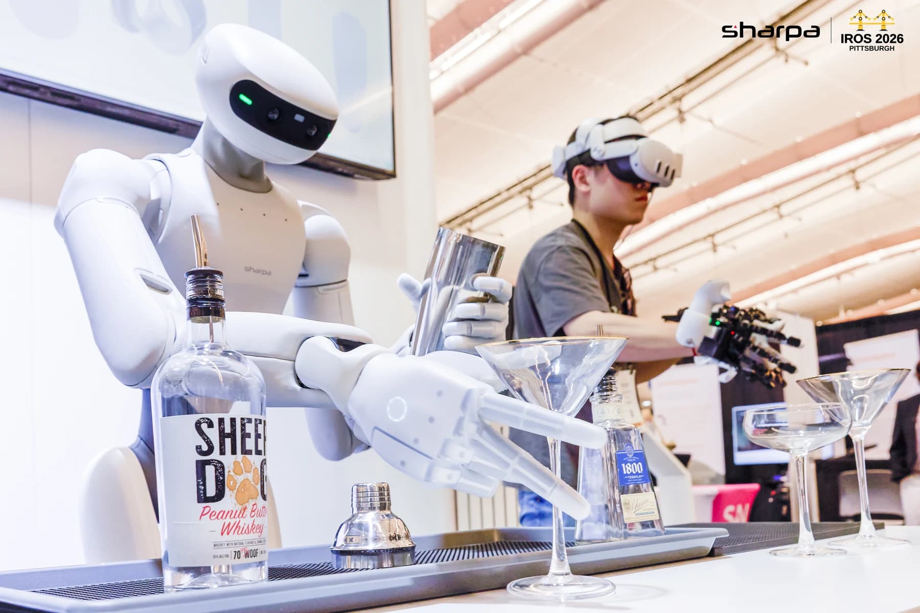 Sharpa robot holding a metal shaker beside an operator wearing a VR headset and exoskeleton gloves at IROS 2026.