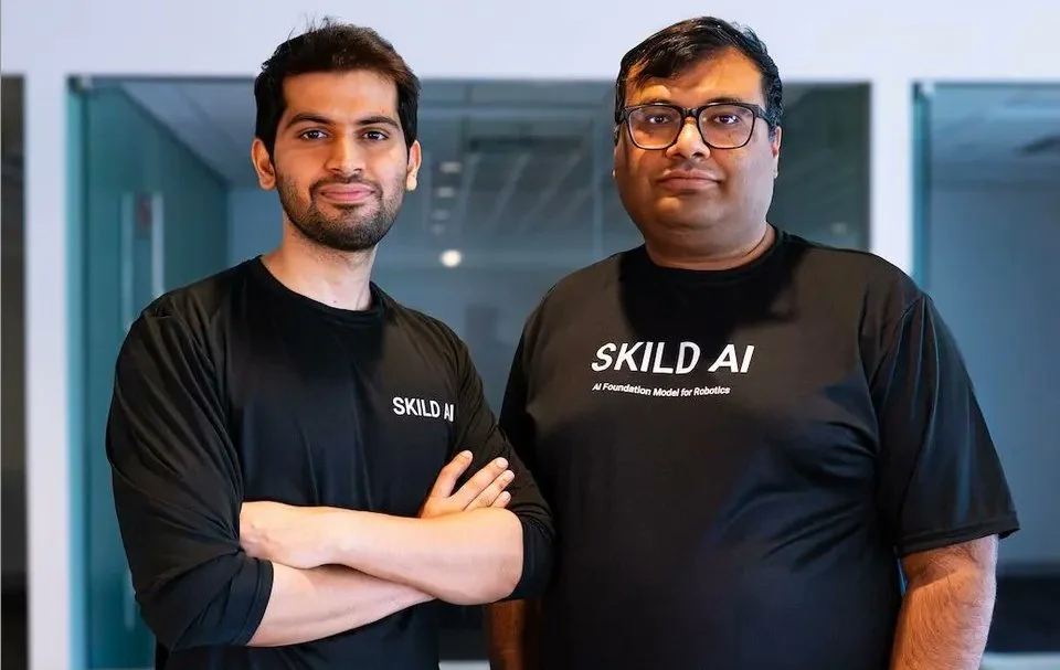 Skild AI co-founders Deepak Pathak and Abhinav Gupta standing side by side wearing black Skild AI branded shirts in an office environment.