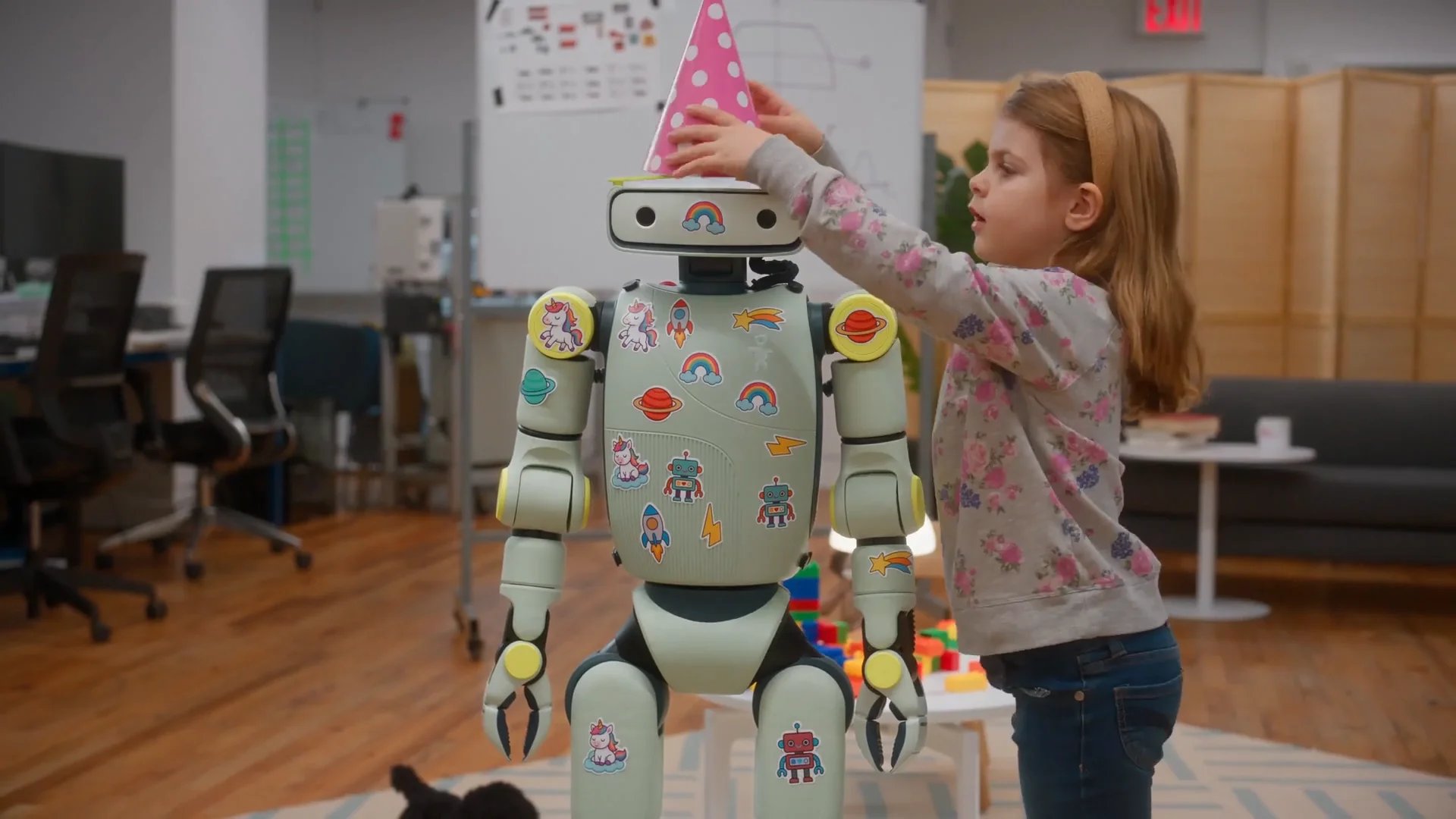 A young girl puts a pink party hat on the Sprout robot, which is decorated with colorful stickers of unicorns, rainbows, and space-themed icons.