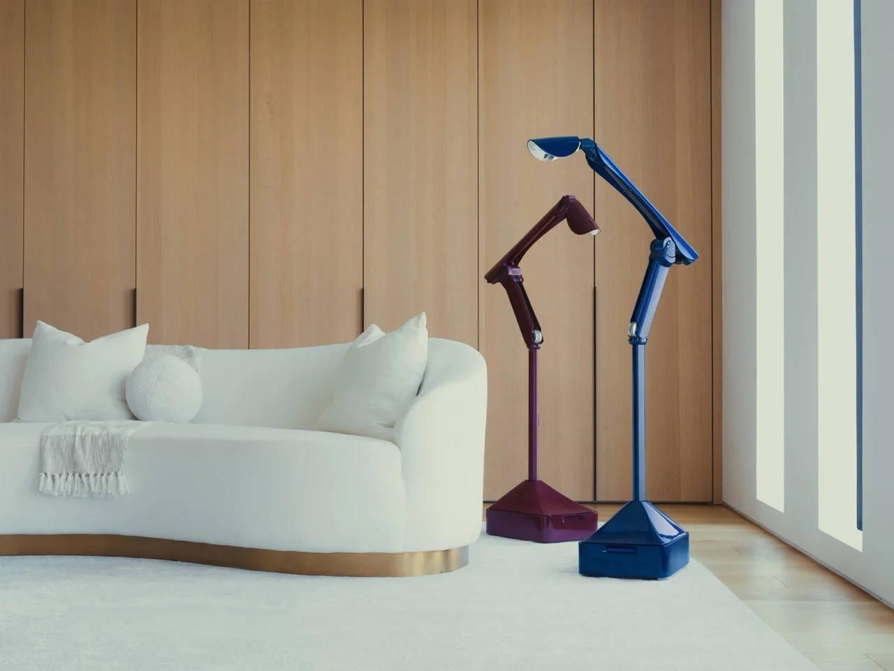 Two Lume robots, finished in high-gloss burgundy and navy blue, standing next to a modern white sofa. The robots feature a slender, articulating neck and a square base, designed to mimic a minimalist floor lamp.