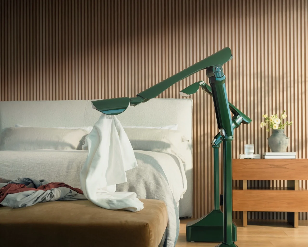 A forest green Lume robot articulating its arm to pick up a white t-shirt from a brown ottoman in a modern bedroom.