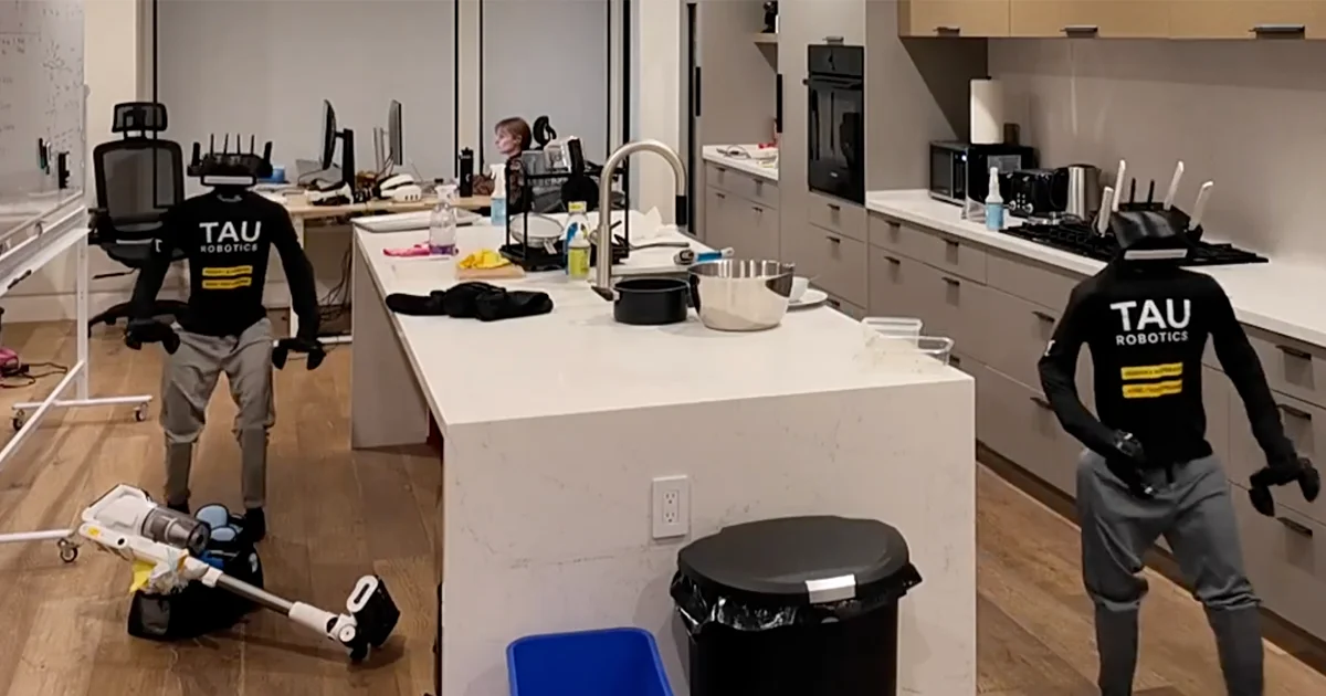 Two humanoid robots wearing black tops with 'TAU ROBOTICS' printed on the chest and grey pants are standing in a modern office kitchen. One robot is positioned near a vacuum cleaner resting on the floor, while the other stands by the kitchen counters. A person is visible sitting at a desk in the background. Both robots feature custom heads equipped with multiple protruding antennas.