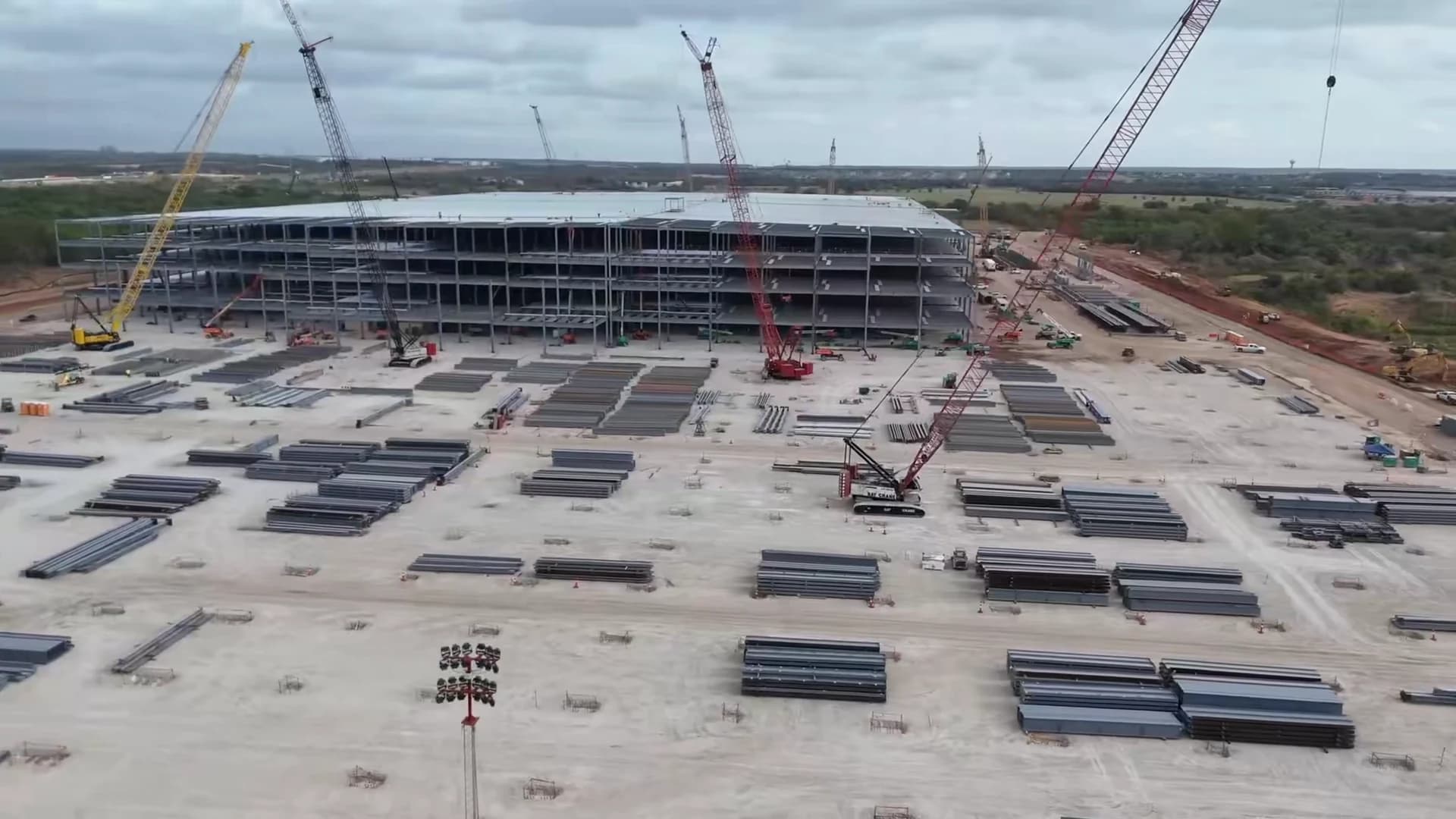 Aerial view of the Optimus factory construction site at Giga Texas, with cranes, a rising steel frame and stacked structural steel.