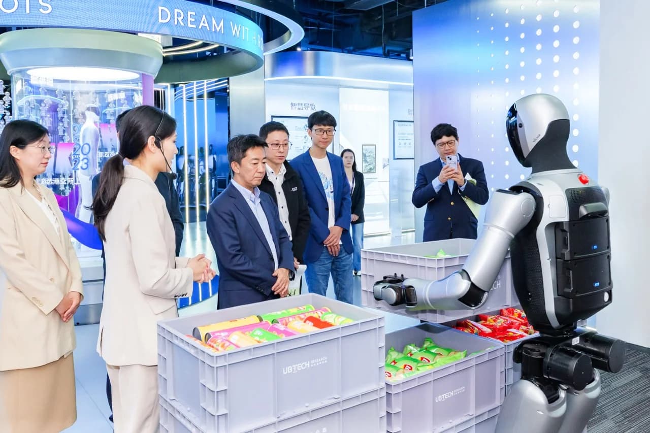 A silver and black Walker S2 humanoid robot reaches into a grey industrial crate labeled "UBTECH RESEARCH" containing packaged snacks while executives from Honda Trading and UBTECH observe the demonstration.