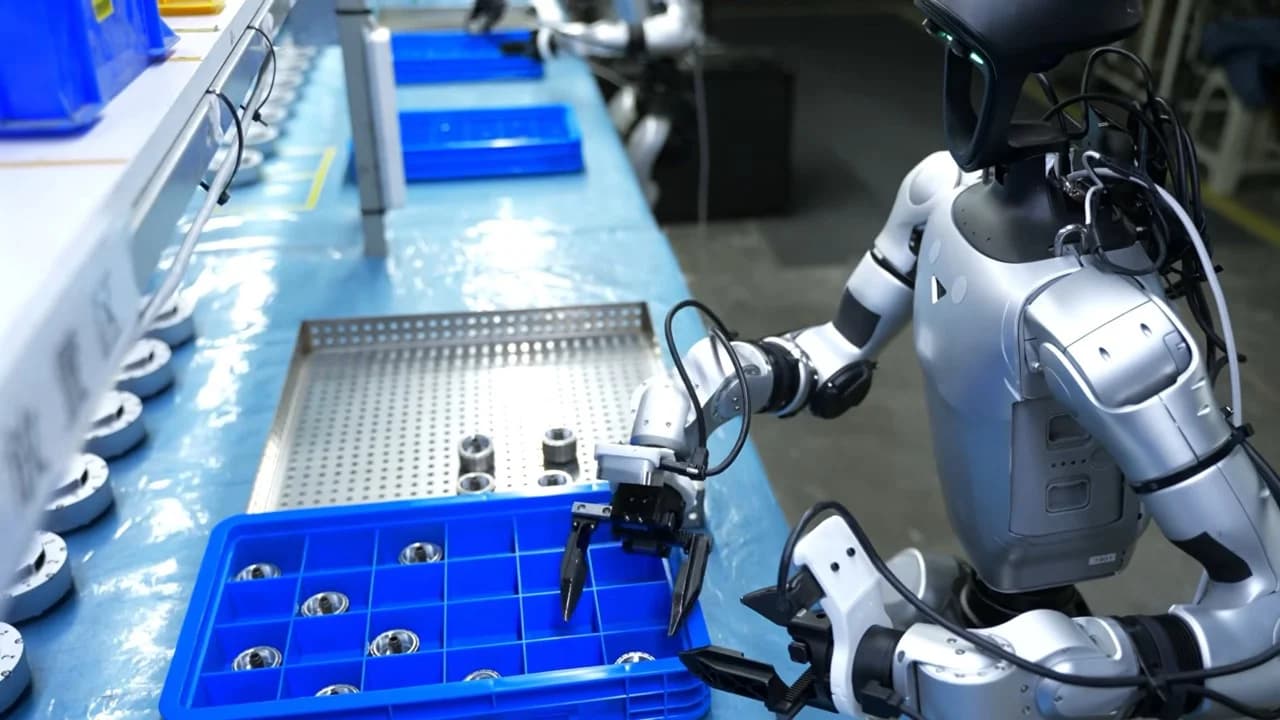 A silver and black Unitree G1 humanoid robot uses its two-fingered grippers to perform precision assembly tasks on small circular components at a factory workbench.