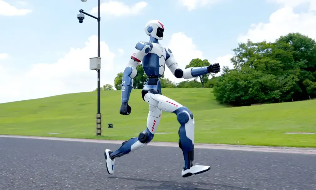 A Unitree R1 humanoid robot running with green trees and bushes in the background