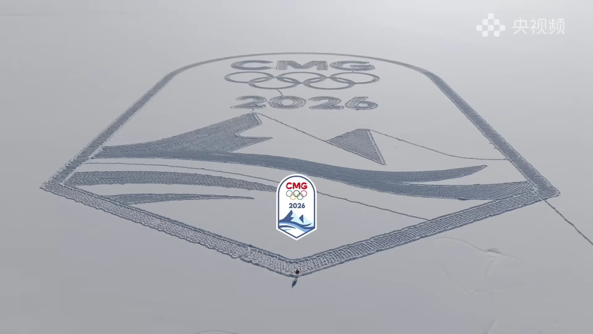 An aerial drone view showing a massive logo for China Media Group's 2026 Winter Olympics coverage, featuring the Olympic rings and "2026," carved into a vast, snowy field.