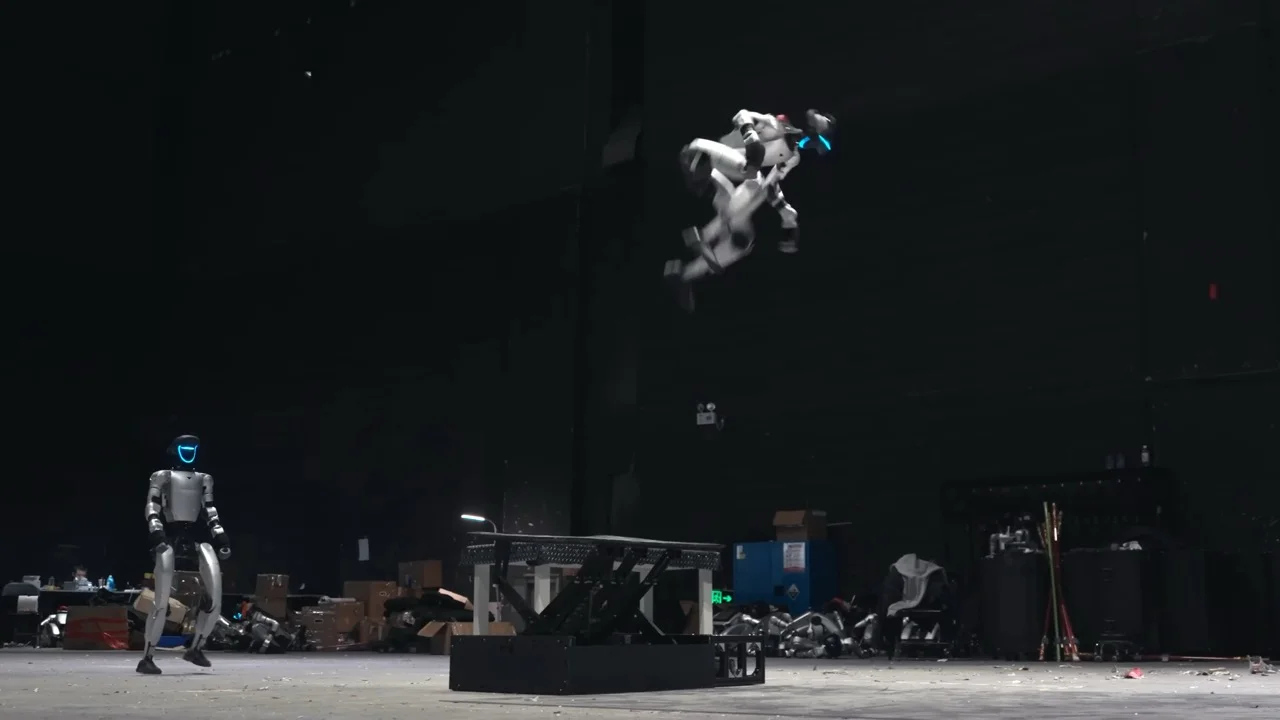 A silver G1 humanoid robot in mid-air, performing a somersault above a black motorized trampoline in a dark rehearsal studio. In the background, another G1 robot stands among equipment and boxes.