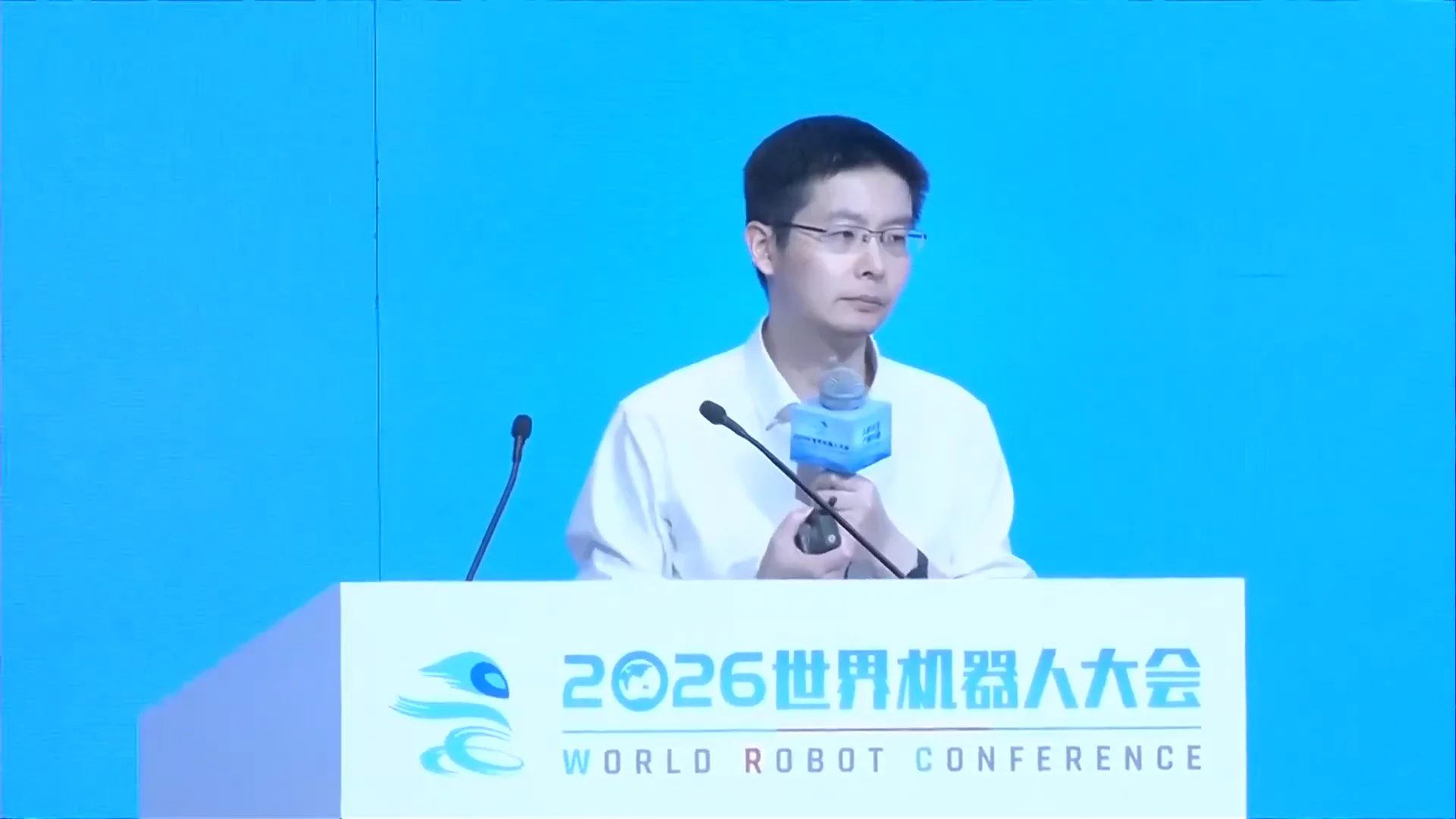 Unitree CEO Wang Xingxing stands at a white podium displaying the 2026 World Robot Conference logo. He is wearing a white collared shirt and glasses, holding a microphone and a presentation clicker against a solid light blue background.