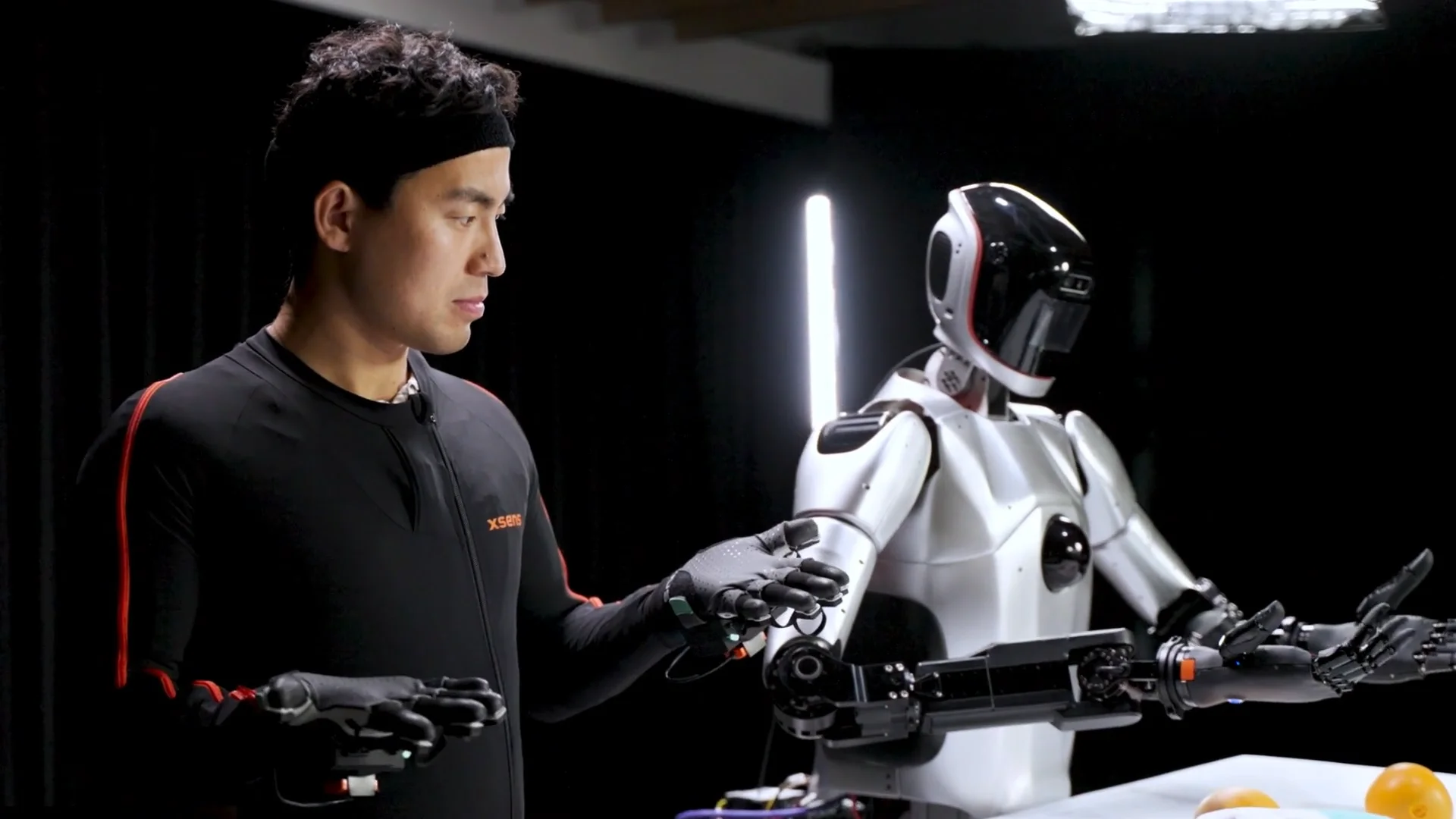 A person wearing a full-body Xsens motion capture suit stands side-by-side with a humanoid robot. The robot is precisely mirroring the person's pose, demonstrating the use of motion capture for teleoperation.